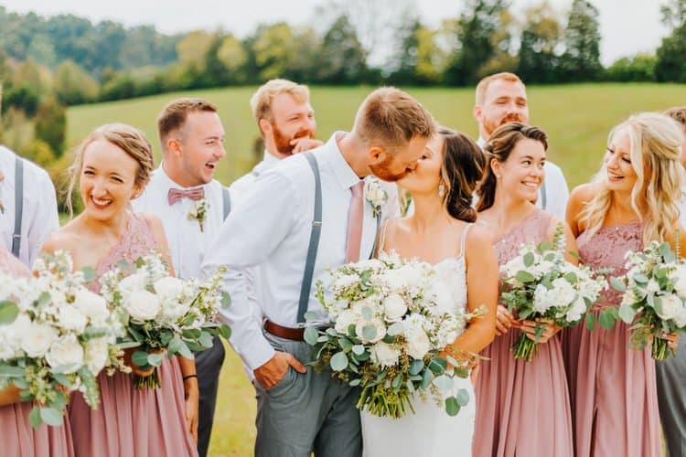 Wedding party outdoors, couple kissing, bridesmaids in dusty rose, groomsmen in grey and white. Green hills background.