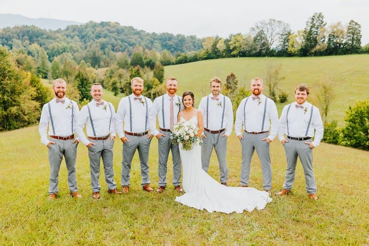 Bride in white dress with bouquet and 7 groomsmen in white shirts, grey pants, and suspenders in a grassy field.