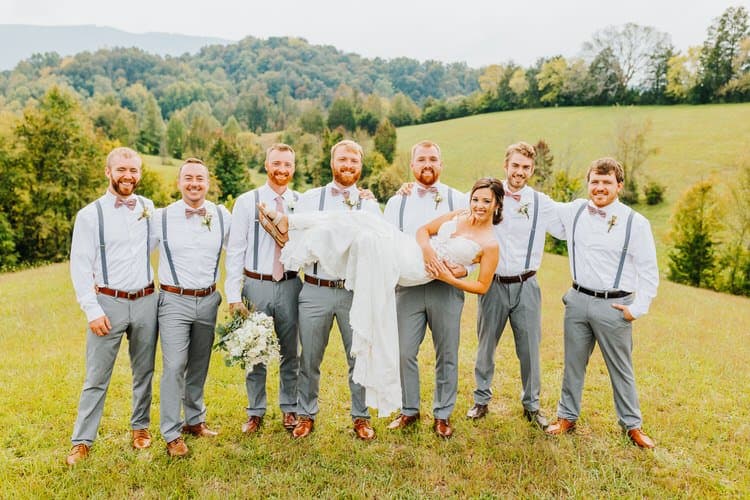 Bride in white dress carried by seven men in white shirts, grey pants, and suspenders in a grassy field with hills.