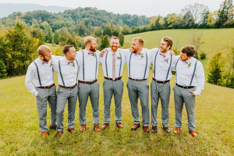 Seven men in white shirts, grey pants, suspenders standing arm-in-arm in a grassy field with mountains at Moose Hollow Lodge.