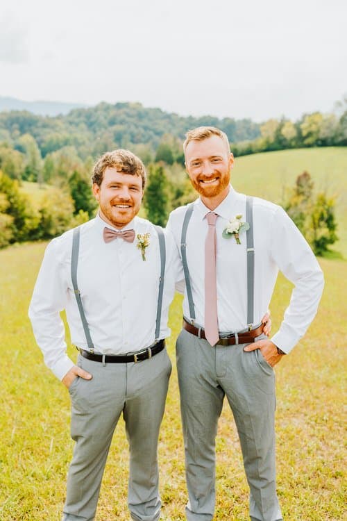 Two men in white shirts, grey suspenders, and light grey pants in a grassy field with rolling green hills in the Smoky...