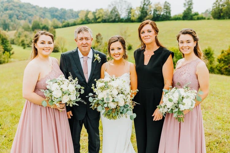 Bride, two women in pink, a man in a suit, and a woman in black stand in a green field with white bouquets.