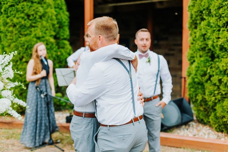 Two men in white shirts and grey pants with suspenders embrace. A woman with a microphone and a man in a bow tie observe....